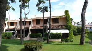 A building with a tiled roof, multiple windows, and a well-maintained lawn with a hedge and trees. - Cozy Villa in a Holiday Village With Pool (Lignano Sabbiadoro)