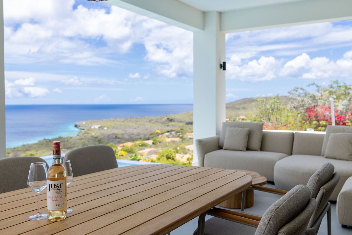 A wooden table with a bottle of wine and glasses, a sofa, and a view of the ocean.