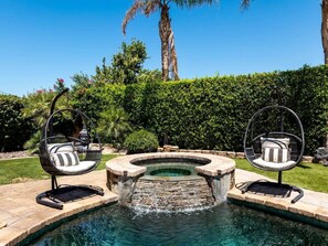 A circular hot tub with a waterfall feature, surrounded by stone steps, with two black swing chairs with white cushions. - Secluded Sanctuary/ Pool/Spa/Casita/BNP/Coachella/Luxury Oasis/Gated Community (La Quinta)
