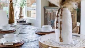 Uma mesa de jantar com um vaso branco, uma janela com cortinas e uma estante com objetos decorativos.