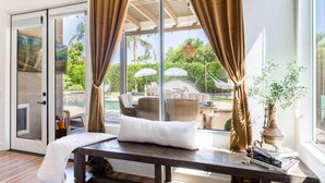 Quarto com uma cama, uma mesa com livros, uma janela com cortinas e vista para móveis de exterior e vegetação.