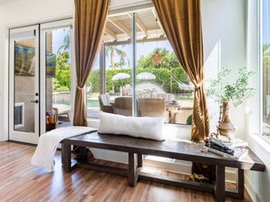 A room with a bed, a table with books, a window with curtains, and a view of outdoor furniture and greenery. - Secluded Sanctuary/ Pool/Spa/Casita/BNP/Coachella/Luxury Oasis/Gated Community (La Quinta)