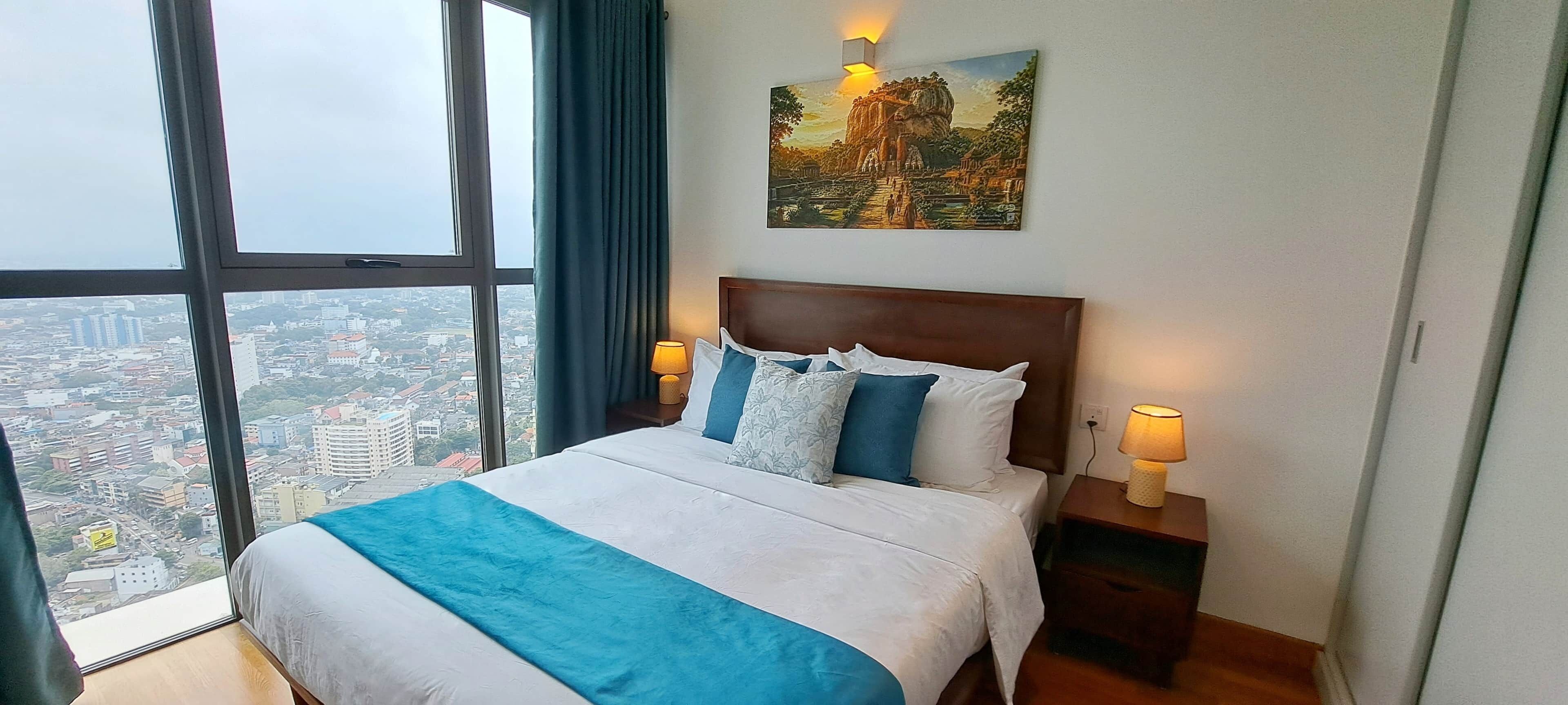 Un cuarto de hotel con un ventanal grande, una cama con ropa de cama azul y blanca, dos lámparas de noche y un cuadro en la pared.