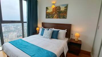 Un cuarto de hotel con un ventanal grande, una cama con ropa de cama azul y blanca, dos lámparas de noche y un cuadro en la pared.
