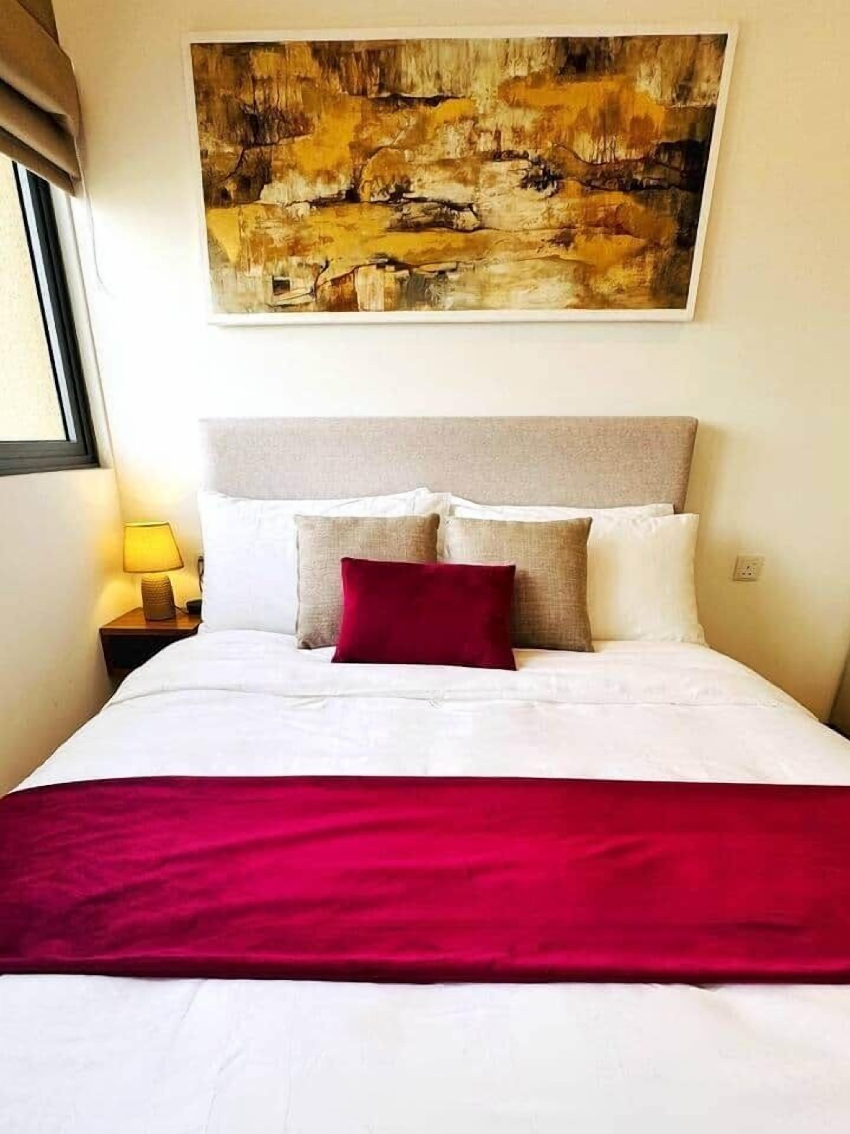 Ein Bett mit weißer Bettwäsche, einem roten Überwurf, einem beige farbenen Kopfteil und einem Gemälde darüber.