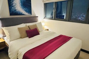 A bed with white bedding and a red throw, a nightstand with a lamp, a cityscape view through a window, and a wave painting on the wall. - 53rd floor TriZen 1BR Apartment - Stunning Views (Colombo)