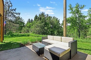 Un espace extérieur aménagé en terrasse, avec des meubles en osier, notamment un canapé et un pouf, le tout se fondant dans un décor de verdure et sous un ciel dégagé.