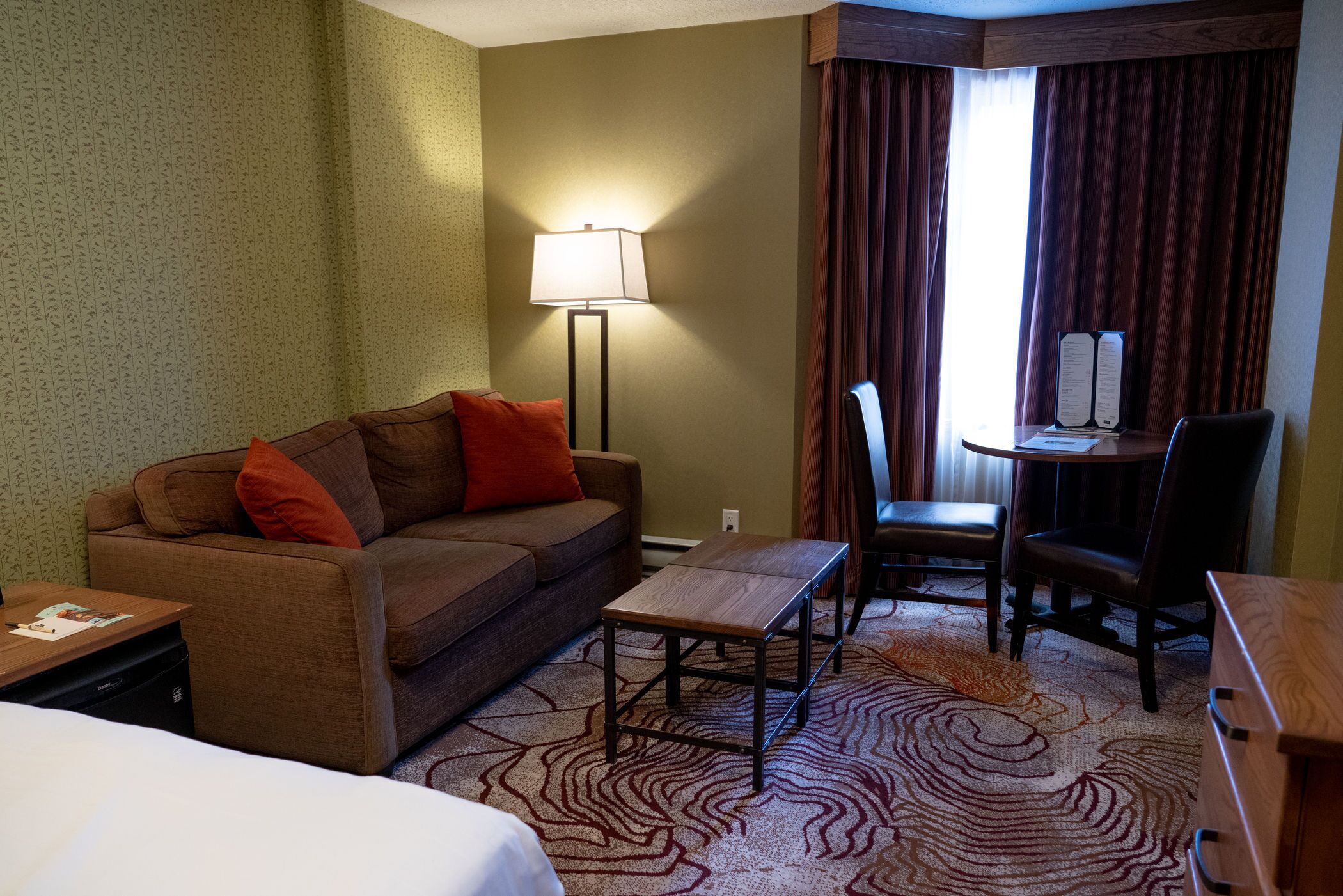 A hotel room with a sofa, a small table, a chair, a lamp, and a view of the outside through a window with curtains.