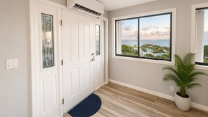 A hallway with a white door, a large window with a view of a coastal area, a potted plant, and a blue mat at the door entrance.