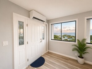 Un couloir avec une porte blanche, une grande fenĂȘtre donnant sur une zone cĂŽtiĂšre, une plante en pot et un tapis bleu devant lâentrĂ©e de la porte.