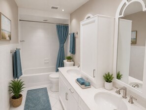 A modern bathroom with a bathtub, toilet, and sink, featuring white tiles, a blue shower curtain, and potted plants.