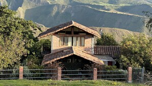 Una cabaña rústica de montaña con techo inclinado, rodeada de abundante vegetación y montañas al fondo.