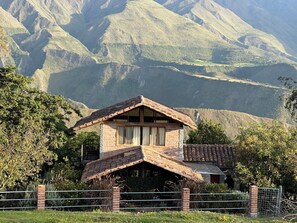 A rustic mountain cabin with a sloped roof, surrounded by lush greenery and mountains in the background.