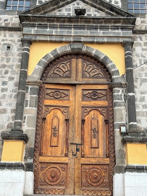 A large wooden double door with intricate carvings, set within a stone archway, flanked by stone columns, and above a yellow wall.