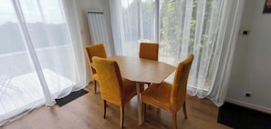 Un comedor con una mesa de madera y cuatro sillas amarillas, ventanas amplias con cortinas translúcidas y un radiador en la pared.