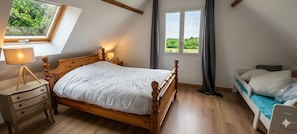 Un dormitorio con cama de madera, una mesita de noche con lámpara, una ventana con cortinas y vistas al exterior con vegetación.