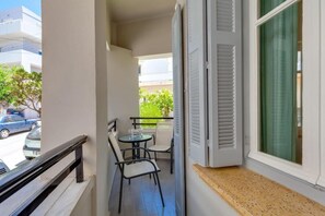 Un balcón con mesa y sillas, una ventana con contraventanas y vistas a la calle y al verdor.