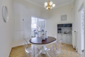 A dining area with a round table, clear chairs, a chandelier, and a kitchen in the background. - Samouil Apartment - Near Beach  (Chania)