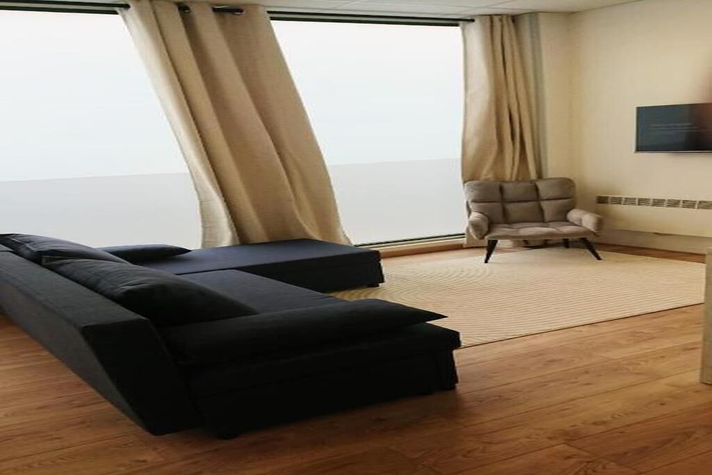 A room with a sofa, a chair, a flat-screen TV, and a view through large windows.