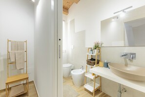 Ett modernt badrum med ett stort handfat, toalett och bidé. Det finns en hylla i trä med handdukar och toalettartiklar.