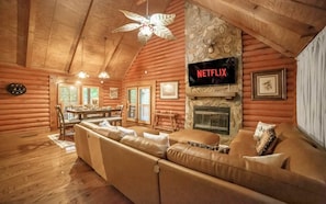 A log cabin interior with a large leather sofa, a stone fireplace, and a mounted TV displaying 'NETFLIX'.