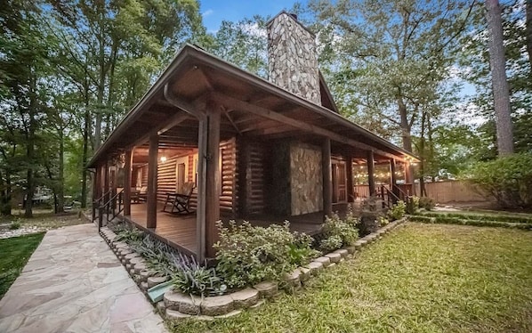 A log cabin with a stone chimney, a porch with seating, and a stone pathway leading to the entrance.
