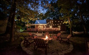 A wooden cabin with a fire pit and outdoor seating, surrounded by trees and string lights.