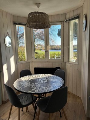A round table with a patterned tablecloth, four black chairs, a woven pendant light, and a view of a lake through large windows. - Le cosy du Green - free pool & golf (Talmont-Saint-Hilaire)
