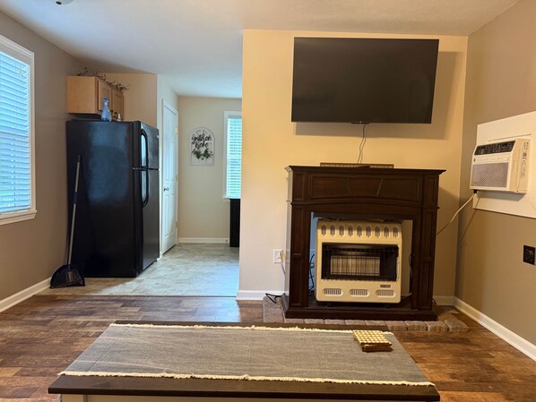 A living room with a fireplace, a television mounted above it, and a kitchen area in the background. - Cabin K- Cozy, Quiet little Cabin near the lake (Nicholas County)