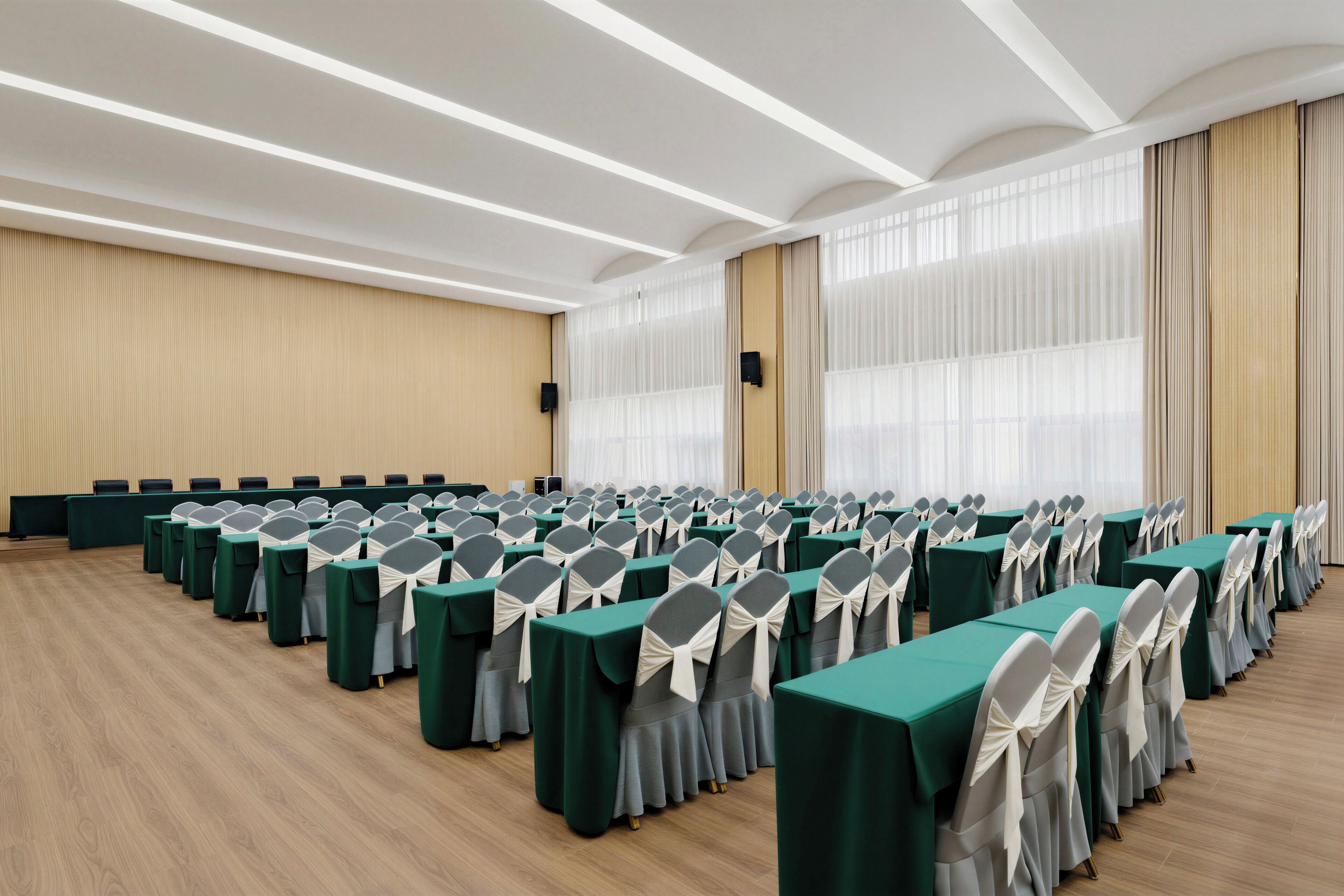 Un salón de conferencias con filas de mesas y sillas, ventanas amplias con cortinas y suelo de madera.