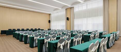 Un salón de conferencias con filas de mesas y sillas, ventanas amplias con cortinas y suelo de madera.