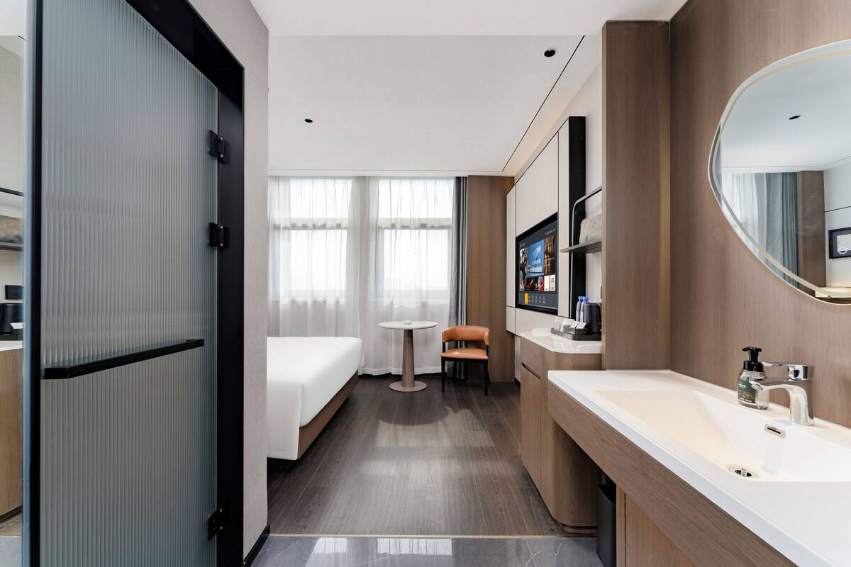 A hotel room with a bed, a small table, a chair, a sink, and a mirror.