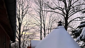 Una cabaña de madera con techo nevado, un porche de madera y una ventana con vista a los árboles circundantes.
