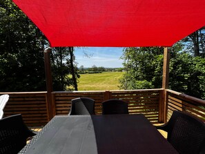 A wooden deck with a table and chairs, a red canopy, and a view of a grassy field. - Prairie cottage with Spa/Pool (Loray)