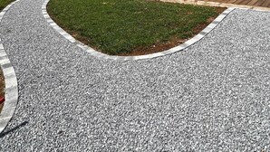 Una cabaña moderna de madera con un patio, rodeada por un camino de grava y vegetación.