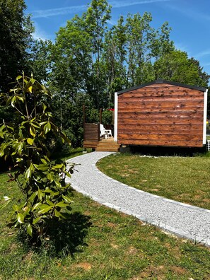 Une cabane en bois, accessible par un chemin de gravier, entourée de verdure et d’arbres.