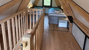 Ett triangulärt sovrum på vinden med synliga träbjälkar, ett fönster, en våningssäng och ett träräcke.
