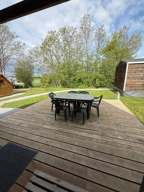 Um deck de madeira com mesa e cadeiras, cercado por vegetação e um céu claro.