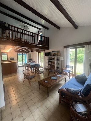 A spacious living area with a balcony, wooden furniture, and a kitchen in the background. - Charming, quiet family home for golf, beach and nature lovers (Moliets-et-Maa)