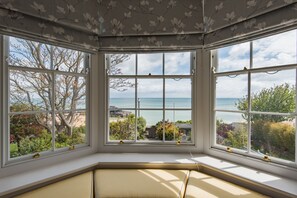 En utåtbuktande fönsterkonstruktion med flera glasrutor, en vadderad bänk och utsikt över havet och den omgivande vegetationen.