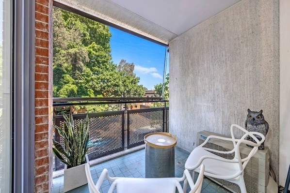 A balcony with a view of trees, a white chair, a table, and a decorative owl statue.