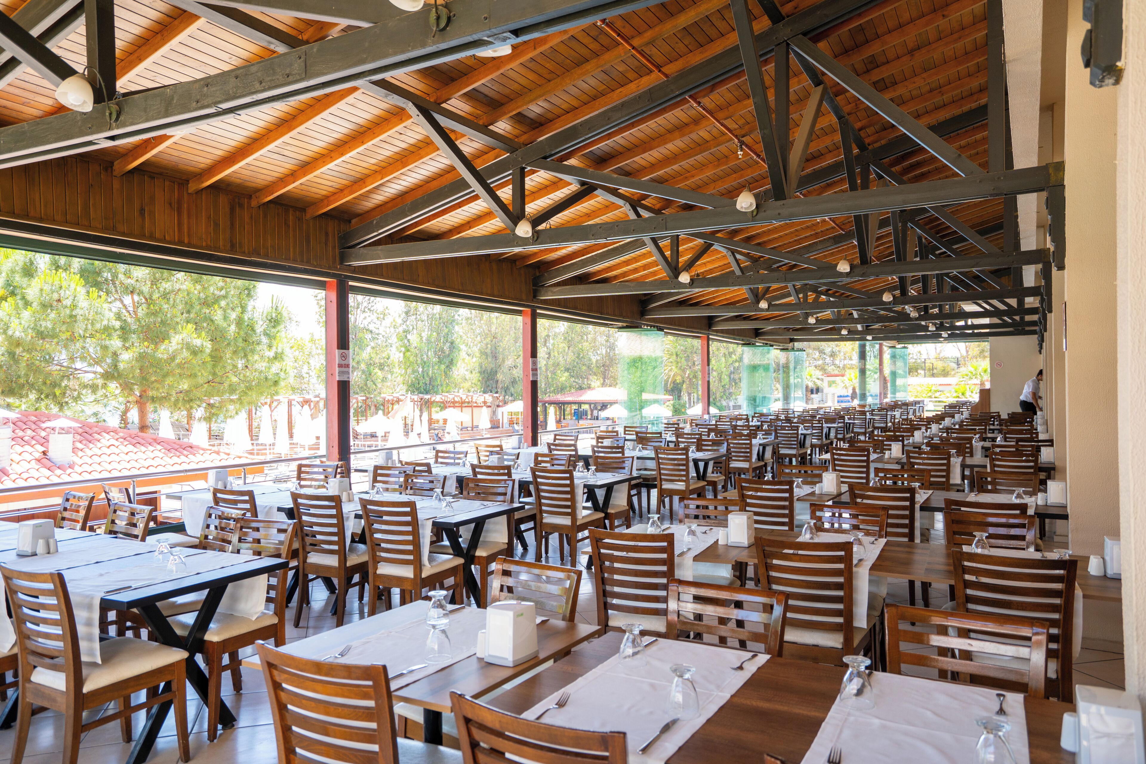 A spacious outdoor dining area with numerous tables and chairs, large windows, and a wooden ceiling structure.