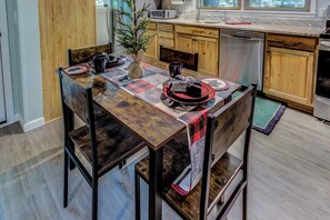 A dining area with a wooden table, chairs, a vase with a plant, and a kitchen in the background. - "Teddy's Rest": Cozy Wooded Cottage in Land Harbor with Lake and Pool Access (Newland)