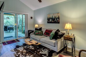 A living room with a sofa, two lamps, a painting, and a view through a sliding door. - "Teddy's Rest": Cozy Wooded Cottage in Land Harbor with Lake and Pool Access (Newland)