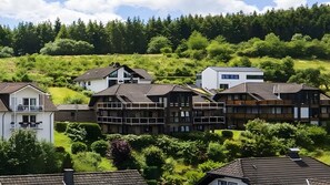 A residential area with houses featuring traditional and modern architecture, surrounded by lush greenery and a forested hillside.