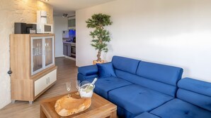 A modern living room with a blue sofa, wooden cabinet, and a potted plant. - Maarhöhe Apartment for 2–6 Guests (54 m²) at Maarberg Resort, Schalkenmehren Lake (Schalkenmehren)