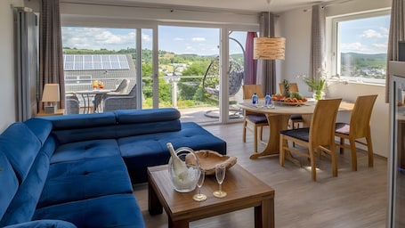 A modern living room with a blue sofa, a dining area with wooden chairs, a table with fruit, and a view of solar panels and greenery outside.. Maarhöhe Apartment for 2–6 Guests (54 m²) at Maarberg Resort, Schalkenmehren Lake