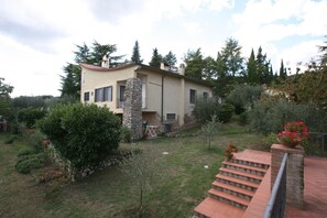 Una casa con facciata in pietra, tetto rosso e un giardino con diverse piante e una scala che conduce a un balcone.
