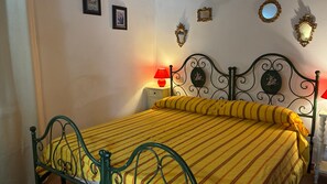 A bed with a metal frame and a yellow striped bedspread, a bedside table with a red lamp, and wall-mounted mirrors and picture frames. - Holiday Home in Villa I Cedri with Pool in Park, 10 Minutes from Siena (Carpineta)
