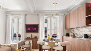 Ein Wohnzimmer mit Sofa, einem Esstisch mit Stühlen, einer Küchenzeile mit Schränken und einem großen Fenster mit Vorhängen.